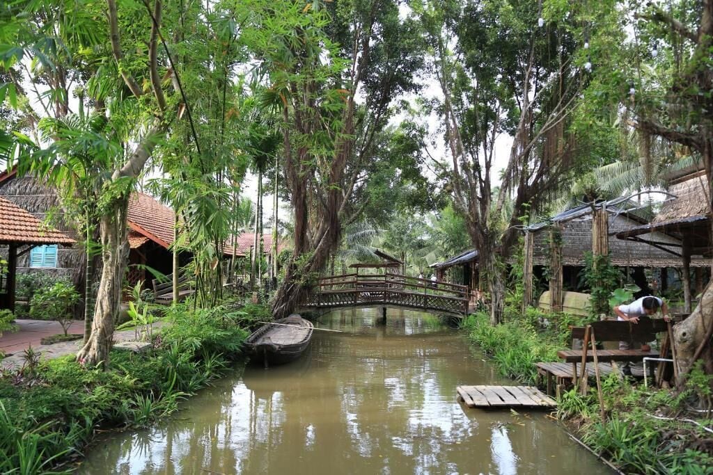 A serene riverside setting with a wooden bridge, lush greenery, and traditional buildings.