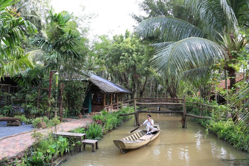 A person in a boat on a calm river surrounded by lush greenery and a wooden bridge.