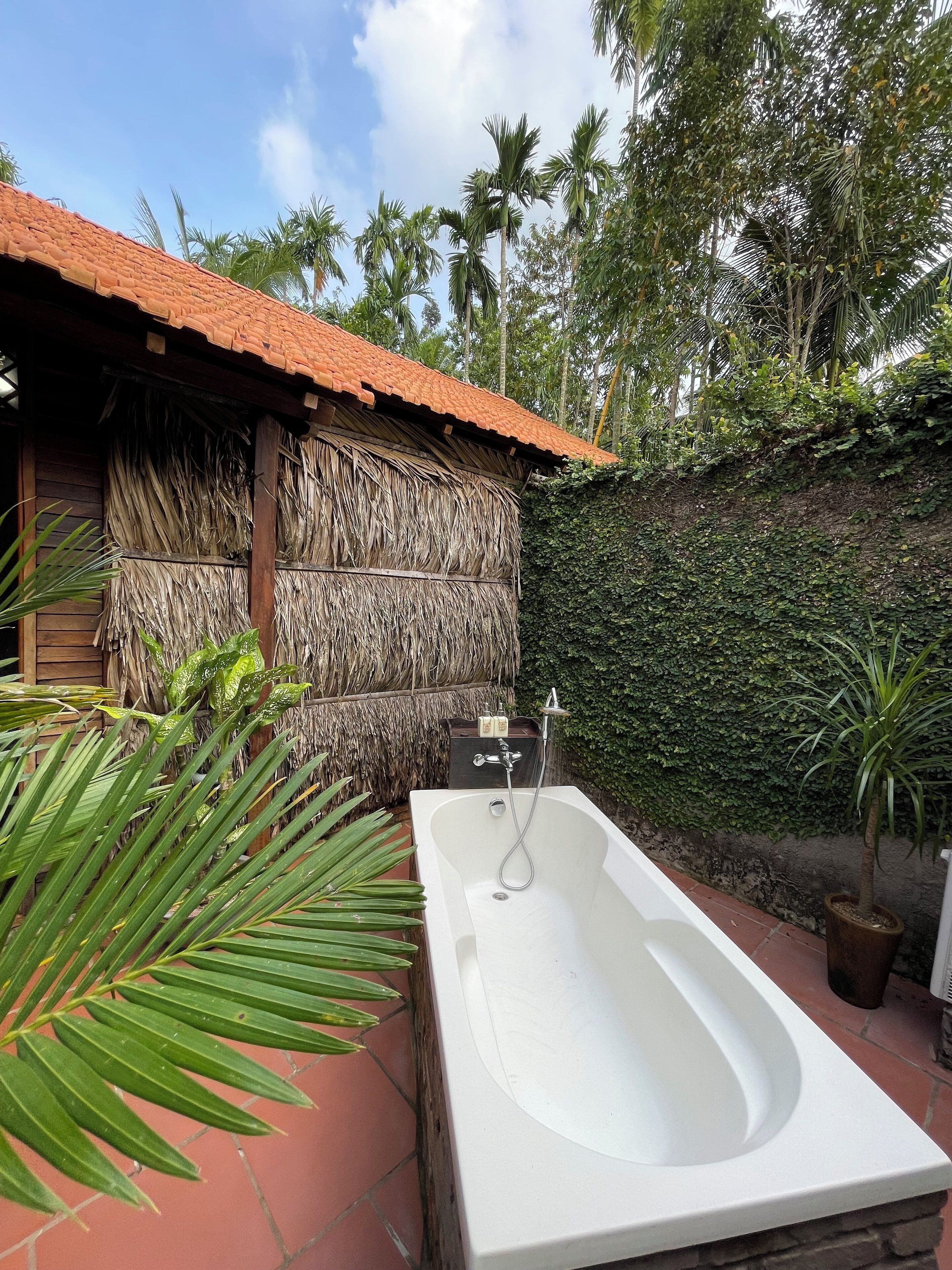 Une baignoire blanche avec une pomme de douche, une structure surmontée d’un toit de chaume et un mur recouvert de verdure.