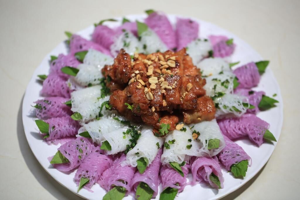 A plate of food with a central meat dish, surrounded by purple and white noodles, garnished with green herbs and peanuts.