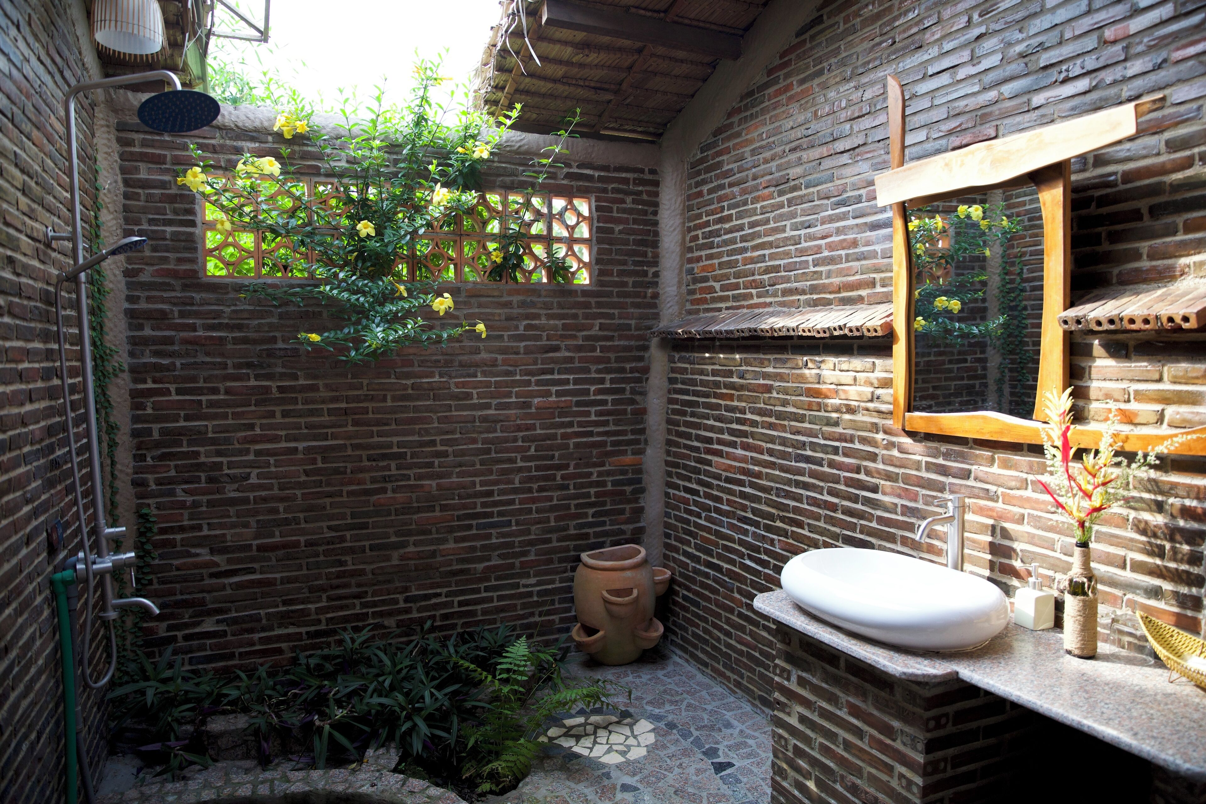 A rustic bathroom with a brick wall, a wooden-framed mirror, a white sink, and a shower area.