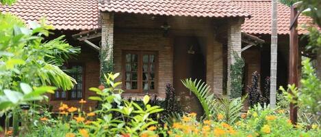 A rustic house with a tiled roof, surrounded by lush greenery and flowering plants.