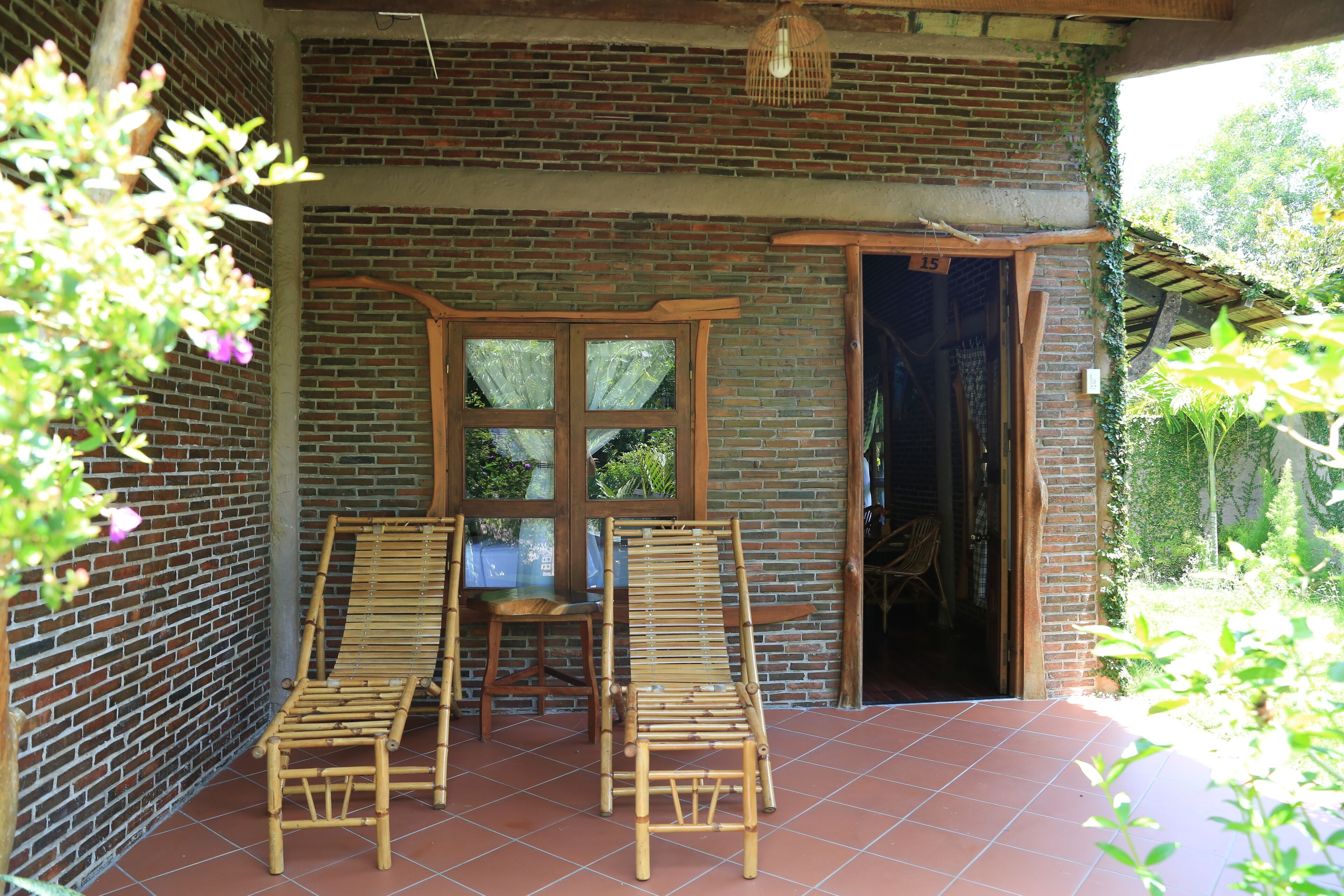 Un patio couvert comprenant deux chaises longues en bambou, une table en bois et un mur en briques avec une fenêtre.