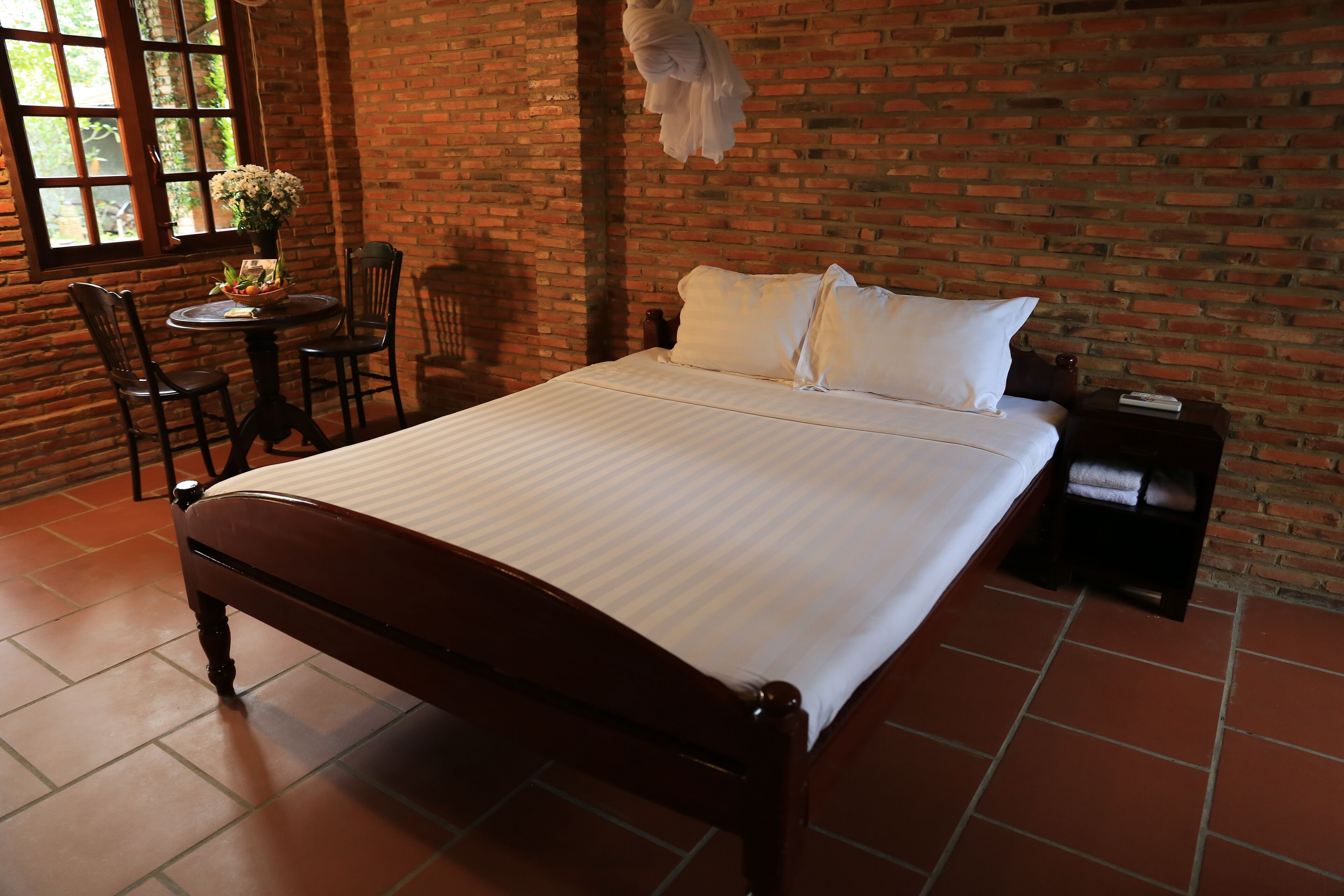 A room with a brick wall, a wooden bed with white bedding, a round table with chairs, and a window with a view of greenery.