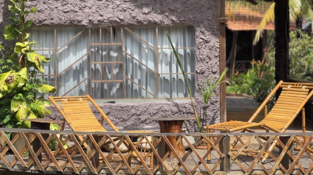 Un edificio de piedra con una ventana, dos sillones de madera y una valla de madera.