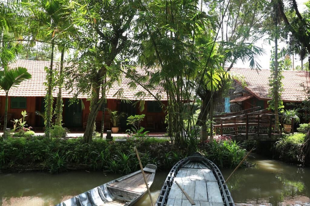 A traditional wooden boat docked at a calm waterway, surrounded by lush greenery and a building with a tiled roof.