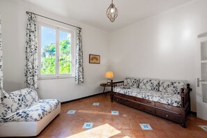 Ein Zimmer mit einer Couch, einem kleinen Tisch mit Lampe, einem Fenster mit Vorhängen und einem Bild an der Wand.