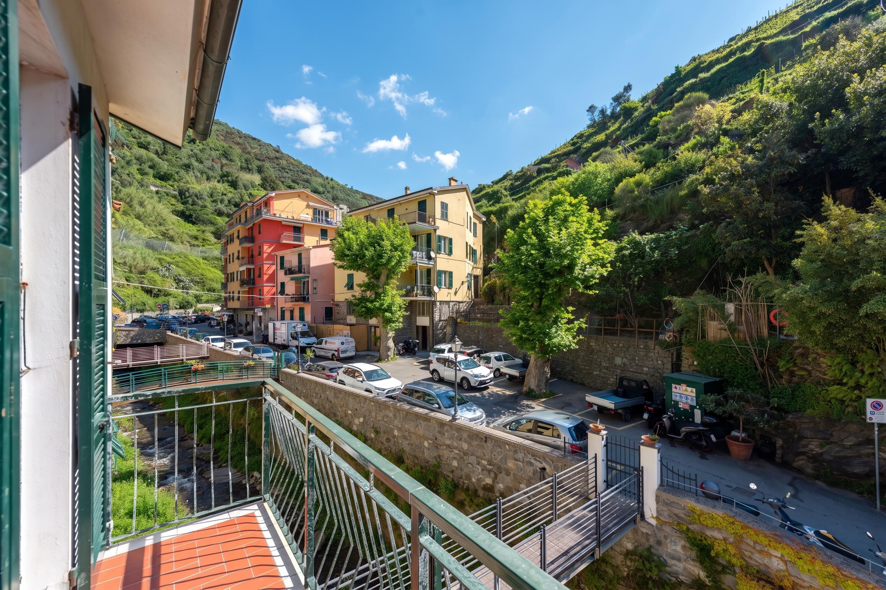 A balcony with a view of a hillside, a parking area with cars, and buildings with balconies.