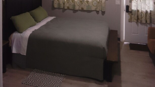 A bedroom with a bed, a bedside table, an air conditioning unit, a window with curtains, and a door. - Charming studio with AC and WiFi in pleasant Gervais. (Gervais)