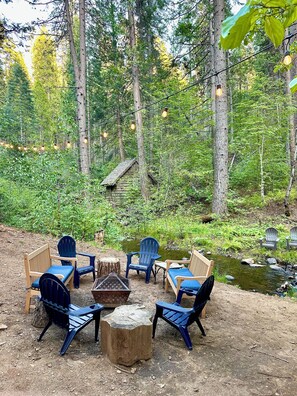 Ein rustikaler Sitzbereich im Freien mit Adirondack-Stühlen und einem Feuerkorb, umgeben von bewaldeter Landschaft.
