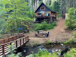 A cabin with a deck, surrounded by a forest, a dirt path, and a small stream. - Creekside Cabin+Fire Pit+Lakes Nearby+Wine Tasting+Bear Valley Ski Resort (Arnold)