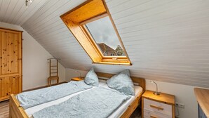 Un dormitorio con techo inclinado, un armario de madera, una cama con colcha estampada, una mesita de noche con lámpara y vistas a través de un tragaluz.