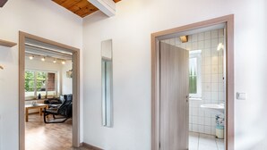 Un pasillo con piso de madera, un espejo, una puerta que da a un baño con lavamanos y ventana, y una sala de estar con sofá y mesa.