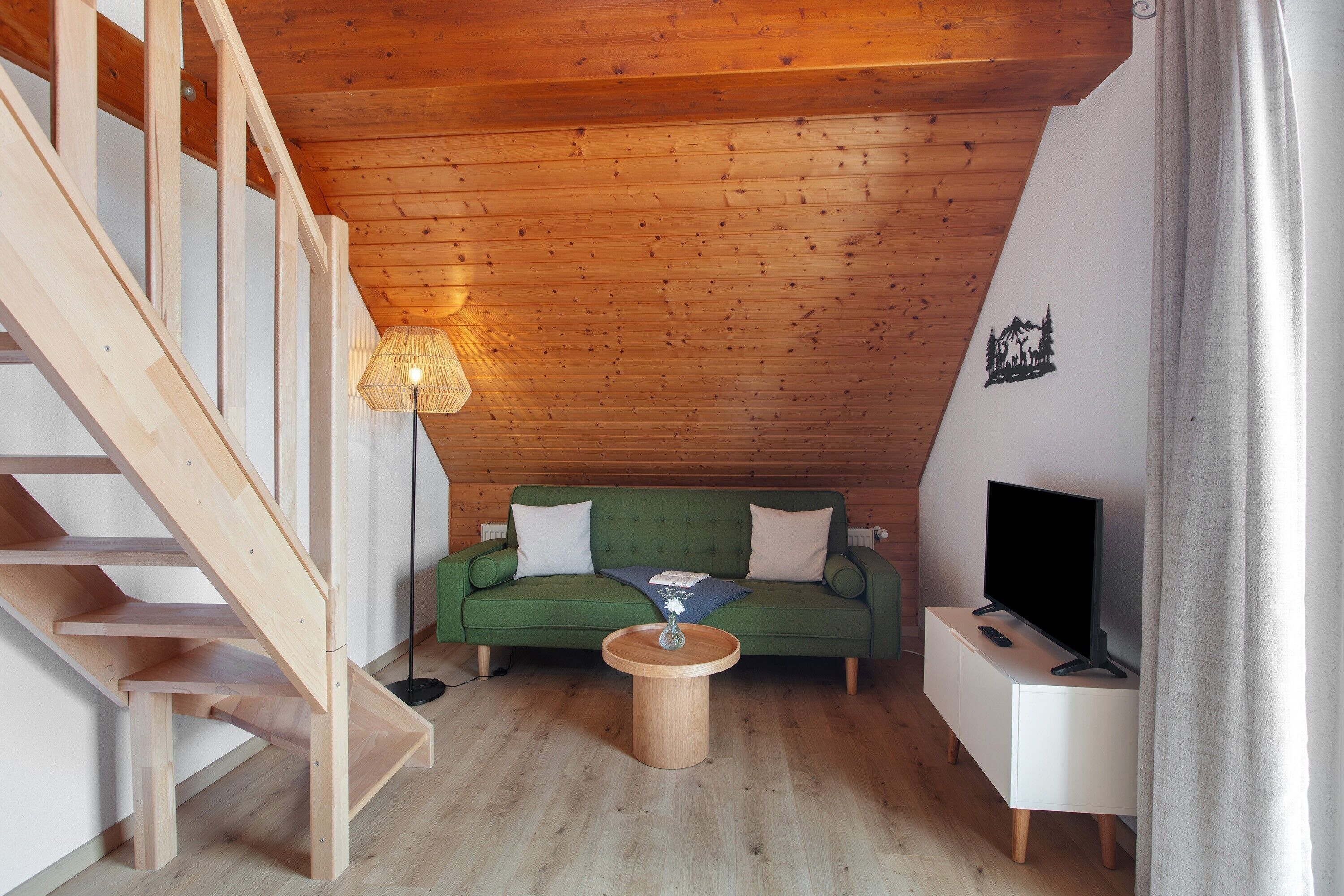 A wooden staircase, a green sofa, a coffee table, a TV on a white stand, and a wall with a forest mural.