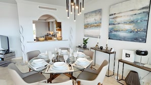 Un coin repas est aménagé avec une table ronde en verre, dressée pour six personnes, et entouré de chaises modernes. On aperçoit une cuisine à travers une arche, et les murs sont ornés de peintures abstraites.