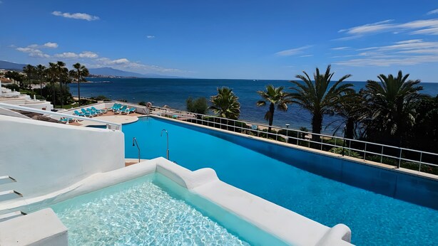 Une piscine à l’eau d’un bleu cristallin, entourée d’espaces blancs aménagés autour du bassin et de palmiers, offrant une vue imprenable sur la côte.