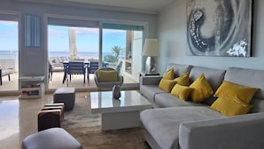 Un salon moderne avec un canapé gris, des coussins jaunes, une table basse et une vue sur la plage grâce à de grandes fenêtres.