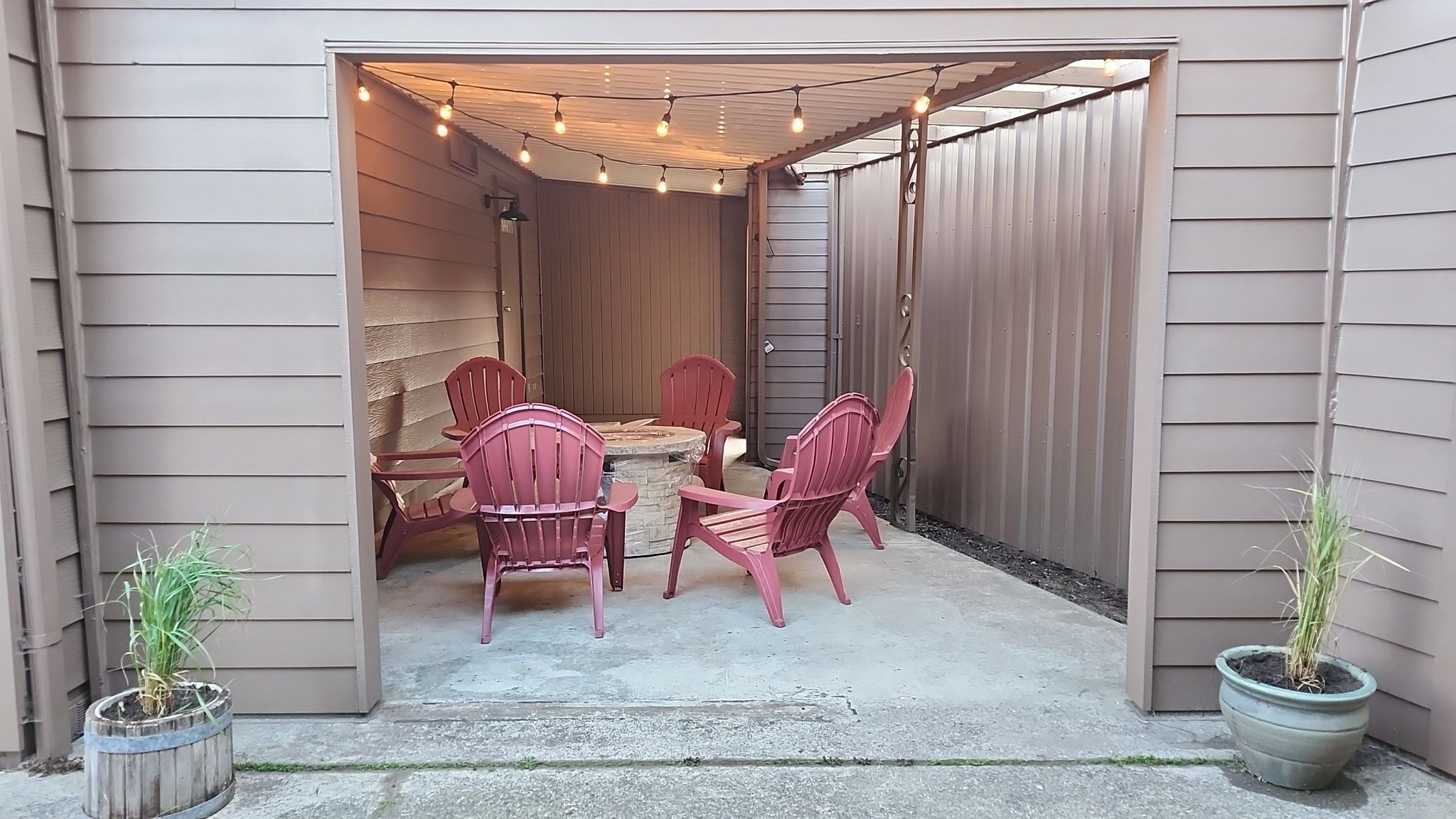 Eine Terrasss mit rundem Tisch, vier Stühlen, Lichterketten darüber und zwei Kübelpflanzen.