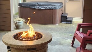 A backyard with a stone fire pit, a hot tub, and red plastic chairs. - Sandy Pacific Hideaway-hot tub, smart tv, woodstove. 4 bedroom home. Florence,OR (Florence)