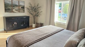 A bedroom with a large bed, a dresser, a potted plant, and a window with curtains. - Sandy Pacific Hideaway-hot tub, smart tv, woodstove. 4 bedroom home. Florence,OR (Florence)