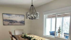 A dining room with a wooden table, chairs, a chandelier, and a large window with a view. - Sandy Pacific Hideaway-hot tub, smart tv, woodstove. 4 bedroom home. Florence,OR (Florence)