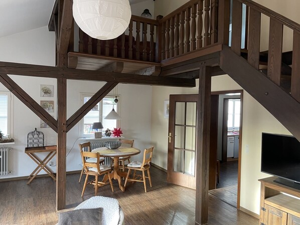 A spacious loft with a high ceiling, exposed wooden beams, a dining area with a round table and chairs, a staircase, and a television on a wooden stand.