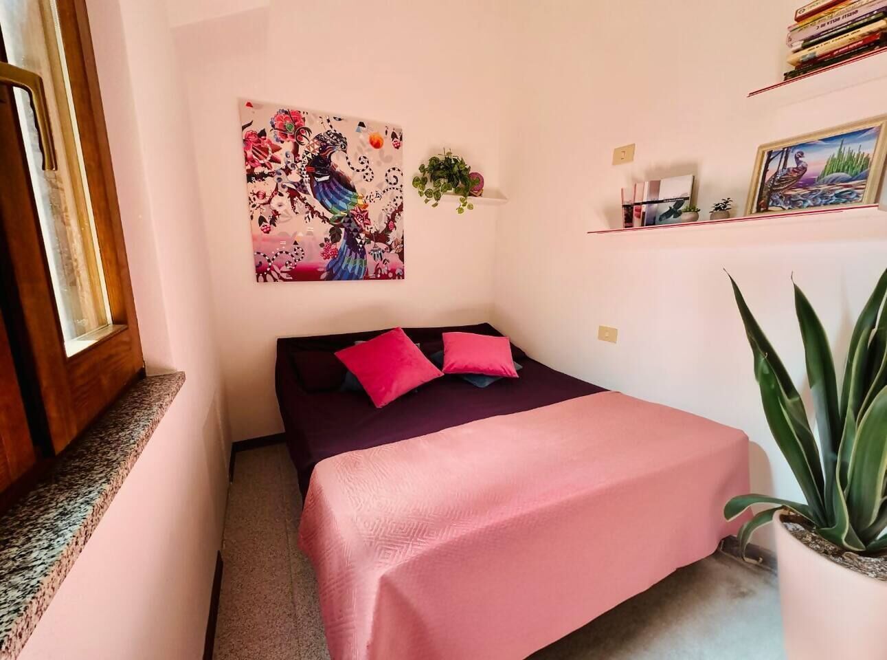 A small, cozy bedroom with a bed, a pink blanket, and a red pillow. There is a painting on the wall and a small plant on a shelf.