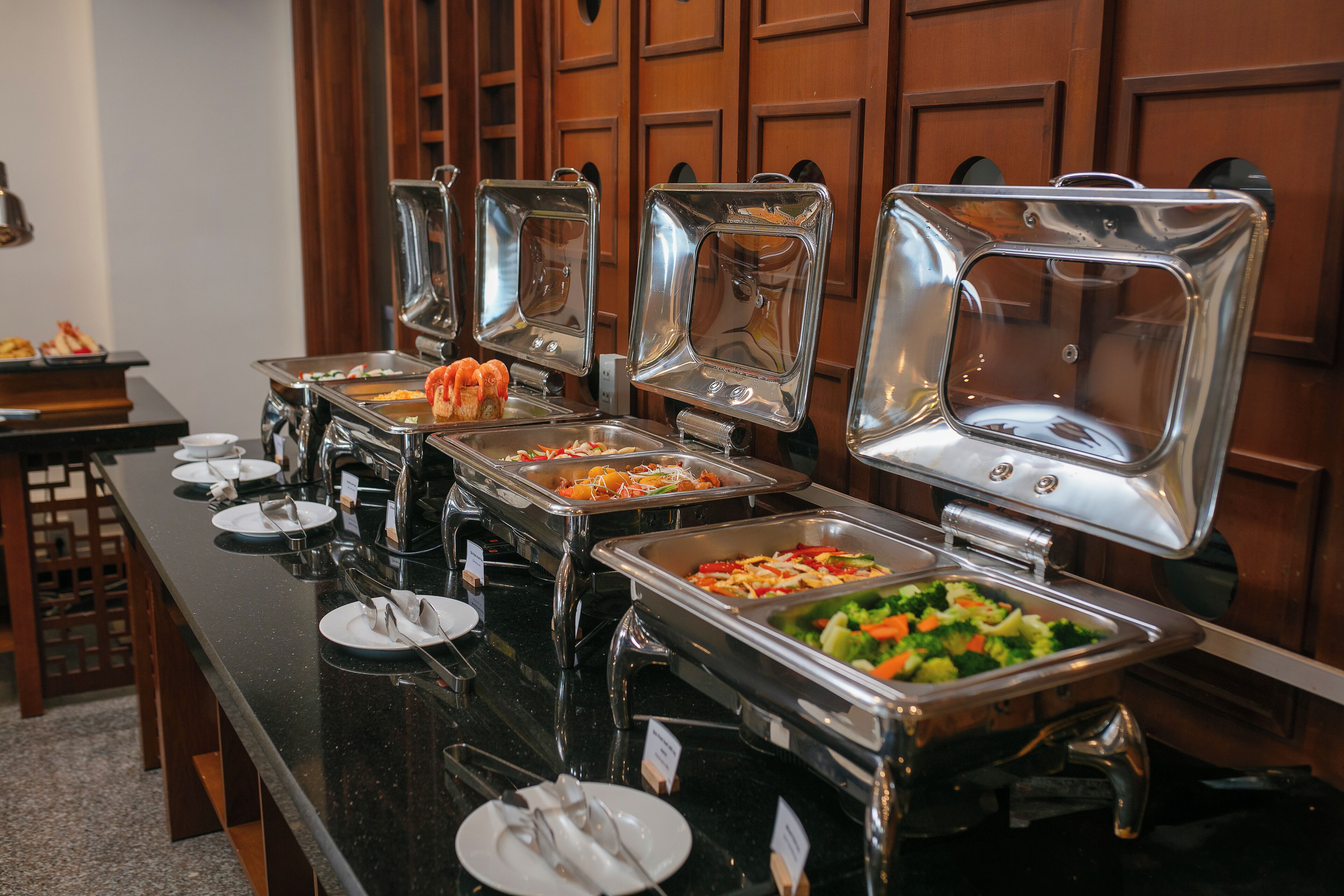 A buffet setup with various food stations, including salads, vegetables, and other dishes, arranged on a long table with metal serving trays.