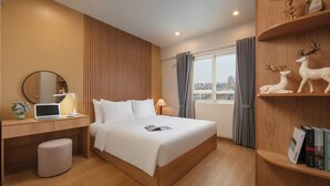 Moderni hotellihuone, jossa on puinen sänky, työpöytä kannettavalla tietokoneella, koriste-esineillä täytetty kirjahylly ja ikkunasta avautuva näkymä kaupunkiin.