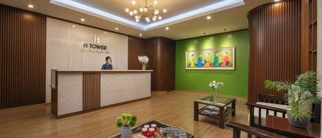 A hotel reception area with a wooden reception desk, a green wall with a painting, a ceiling light fixture, and a potted plant.