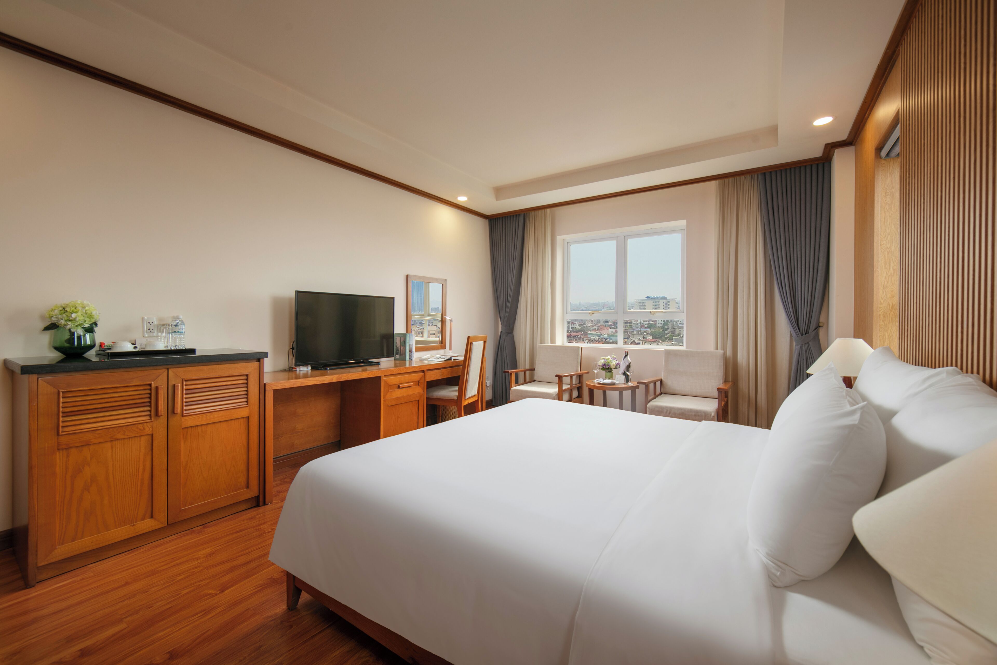 Habitación de hotel con una cama grande, un escritorio con televisor, una mesita con lámpara y vistas a la ciudad por la ventana.