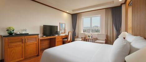 Habitación de hotel con una cama grande, un escritorio con televisor, una mesita con lámpara y vistas a la ciudad por la ventana.