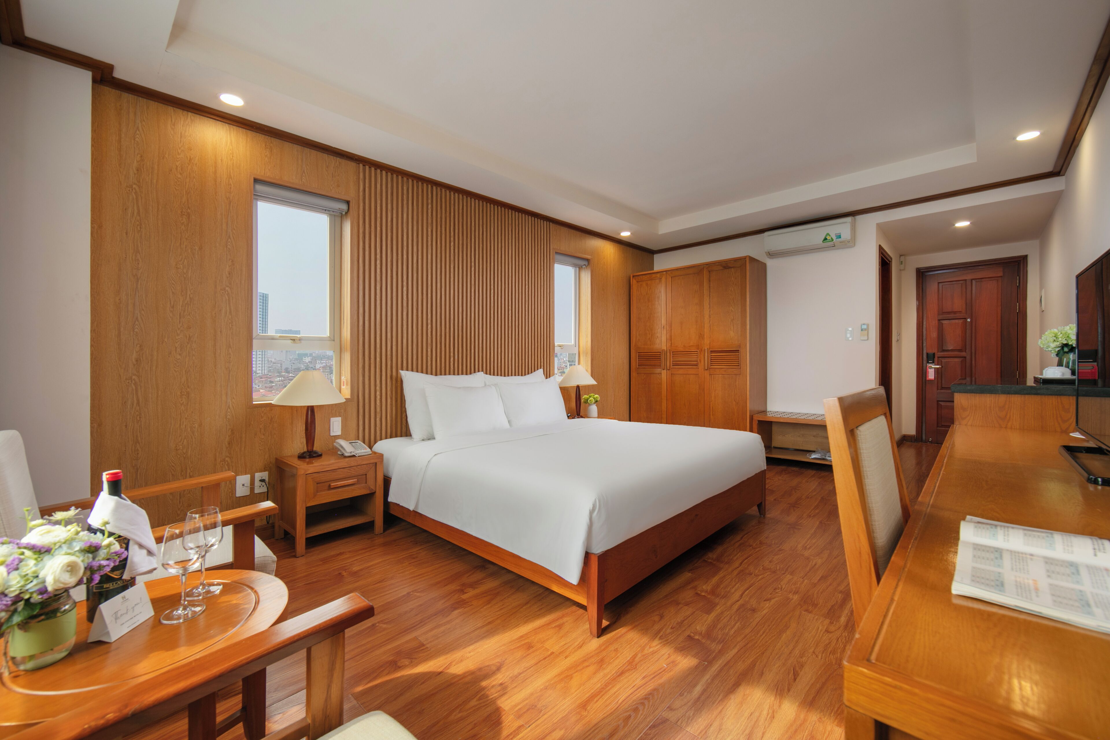 A hotel room with a large bed, wooden flooring, a desk with a chair, a small table with a vase of flowers, and a window with a city view.