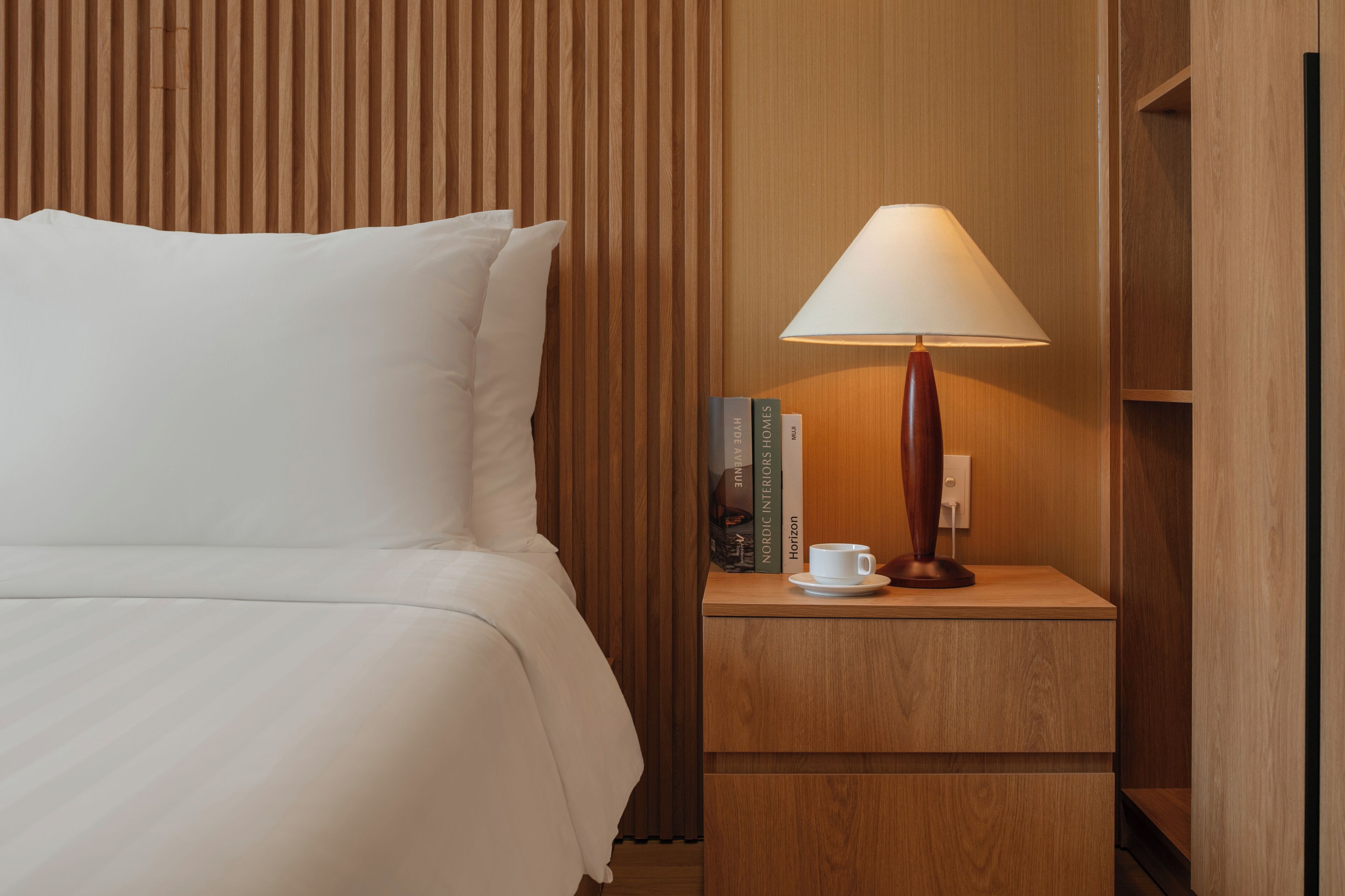 A neatly made bed with white linens, a wooden nightstand with a lamp, a cup, and a few books.