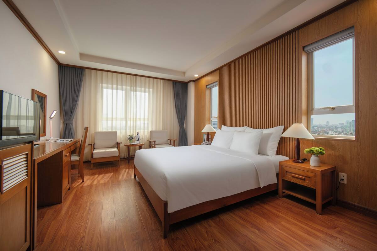 A hotel room with a wooden floor, a large bed with white linens, a desk with a television, a chair, and a nightstand with a lamp and a plant.