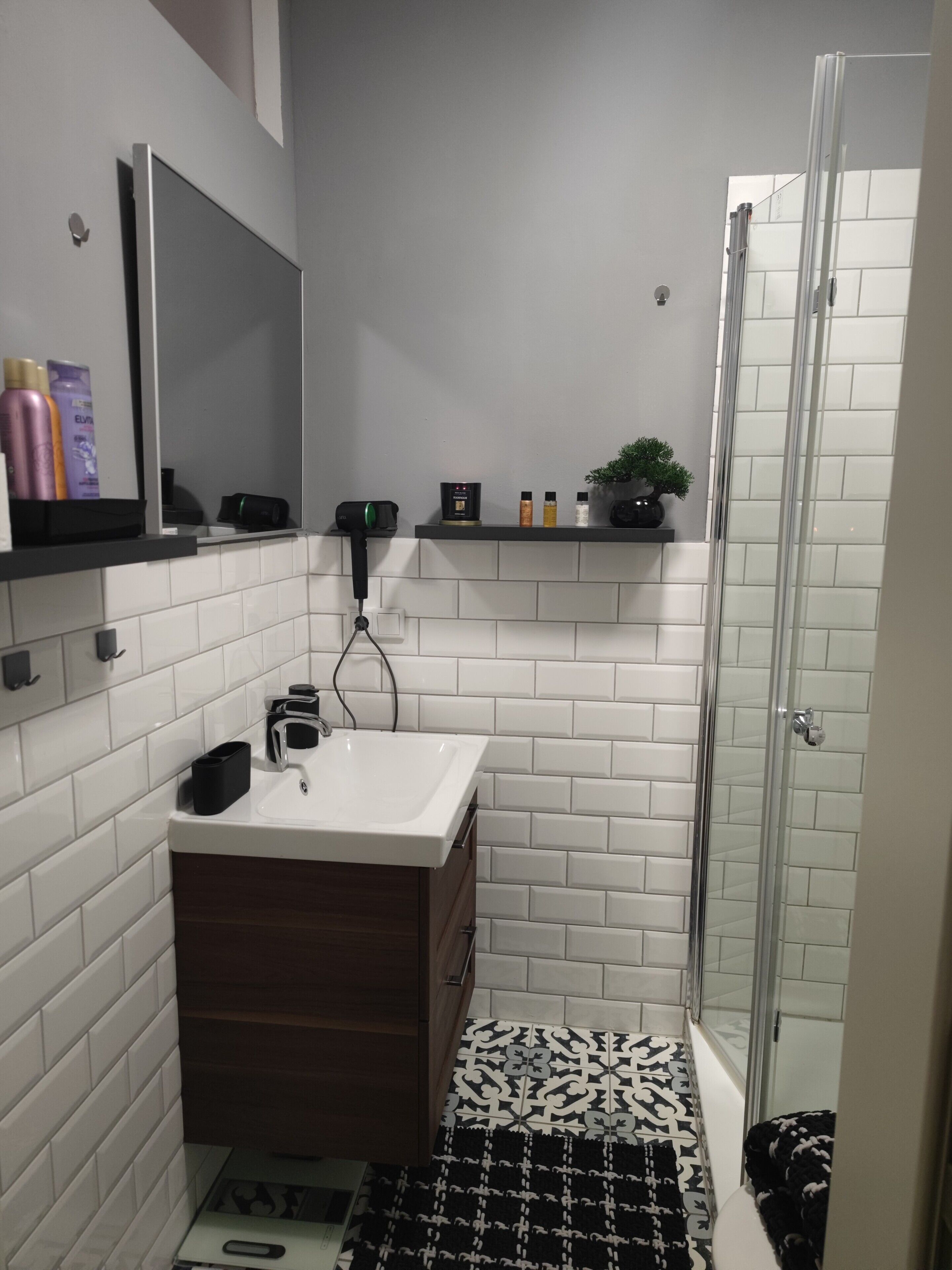 Un baño con pared de azulejos blancos, un lavabo, un espejo, una ducha y una alfombra estampada.