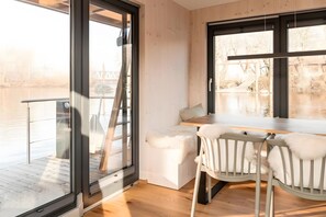 Une chambre moderne avec une porte vitrée donnant sur un balcon, une échelle en bois, un banc blanc avec coussin et une table avec des chaises.