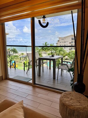 A room with a large sliding glass door leading to a balcony with a pool, outdoor furniture, and a view of the beach and buildings.