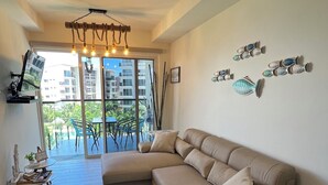 A modern living room with a large L-shaped beige sofa, a small round ottoman, a flat-screen TV on a stand, and a dining area with a table and chairs. - Beachfront Condo | Caracol Beach, Punta Chame | Pools & Ocean Views (Chame)