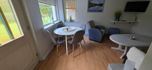 Une pièce comprenant une table de salle à manger ronde, quatre chaises, une petite table ronde avec un saladier, un téléviseur fixé au mur et une fenêtre avec des stores.