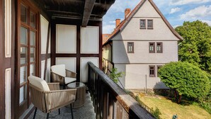 Un balcon avec des fauteuils en osier et une table, une balustrade en bois, et une vue sur un bâtiment blanc doté d’un toit à deux versants.