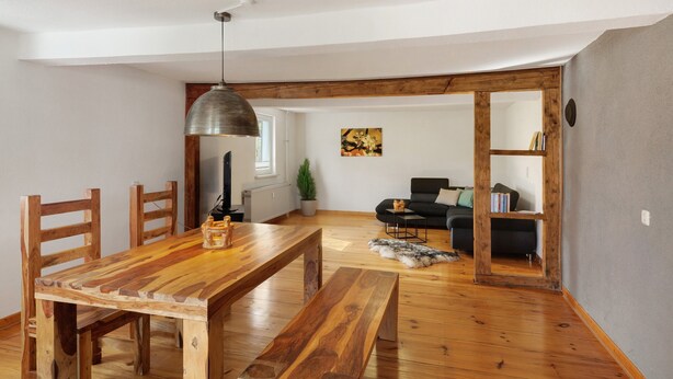 Un coin repas avec une table et des chaises en bois, un luminaire suspendu et une vue sur un salon comprenant un canapé et une bibliothèque.