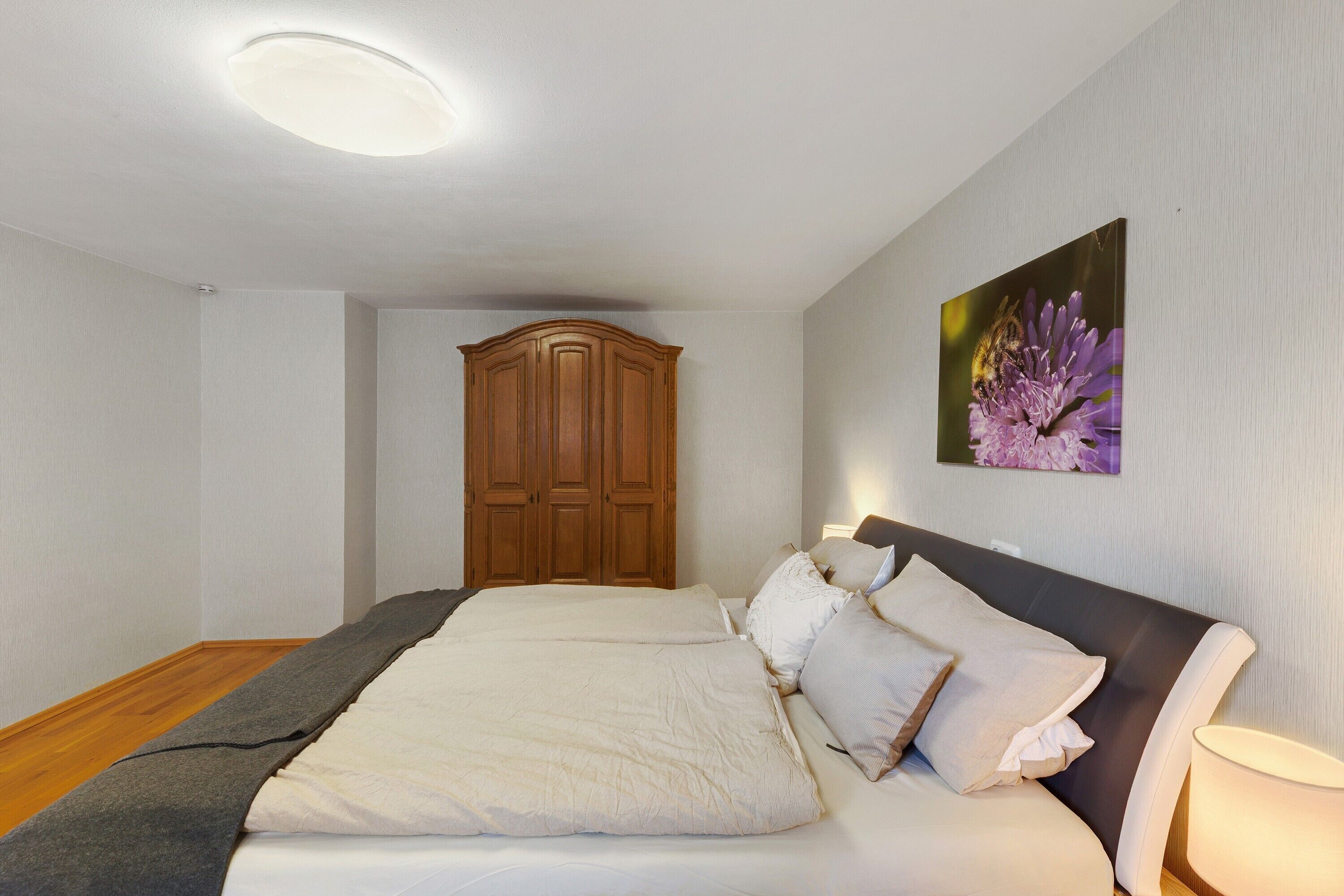 A bedroom with a large bed, a wooden wardrobe, and a wall-mounted lamp.