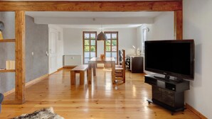 Un salon spacieux doté d’un plancher en bois, d’une table à manger, de bancs, d’un téléviseur posé sur un meuble et d’une grande fenêtre offrant une vue sur la verdure.