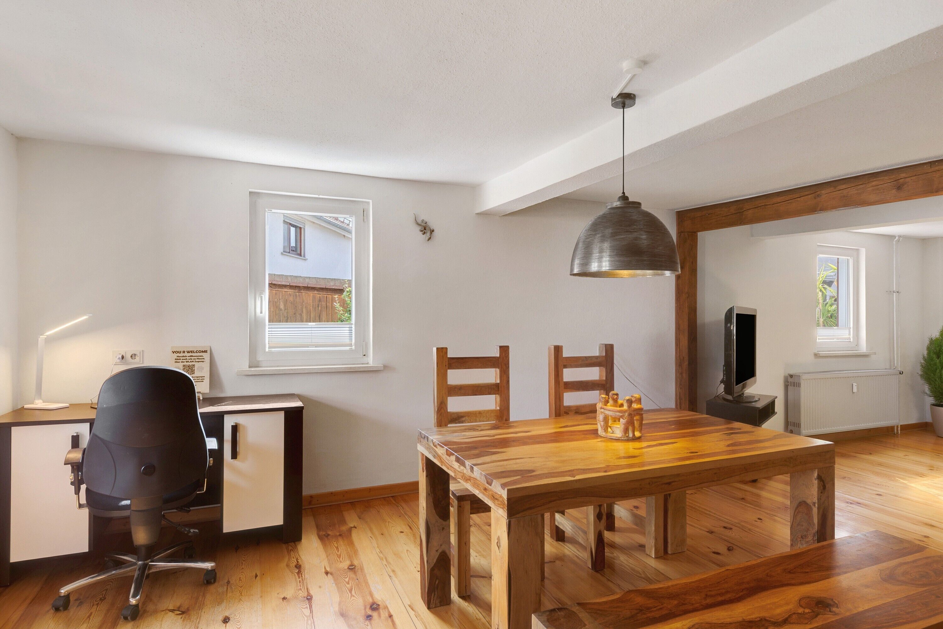 A room with a wooden dining table, a black office chair, a desk with a computer, and a television.