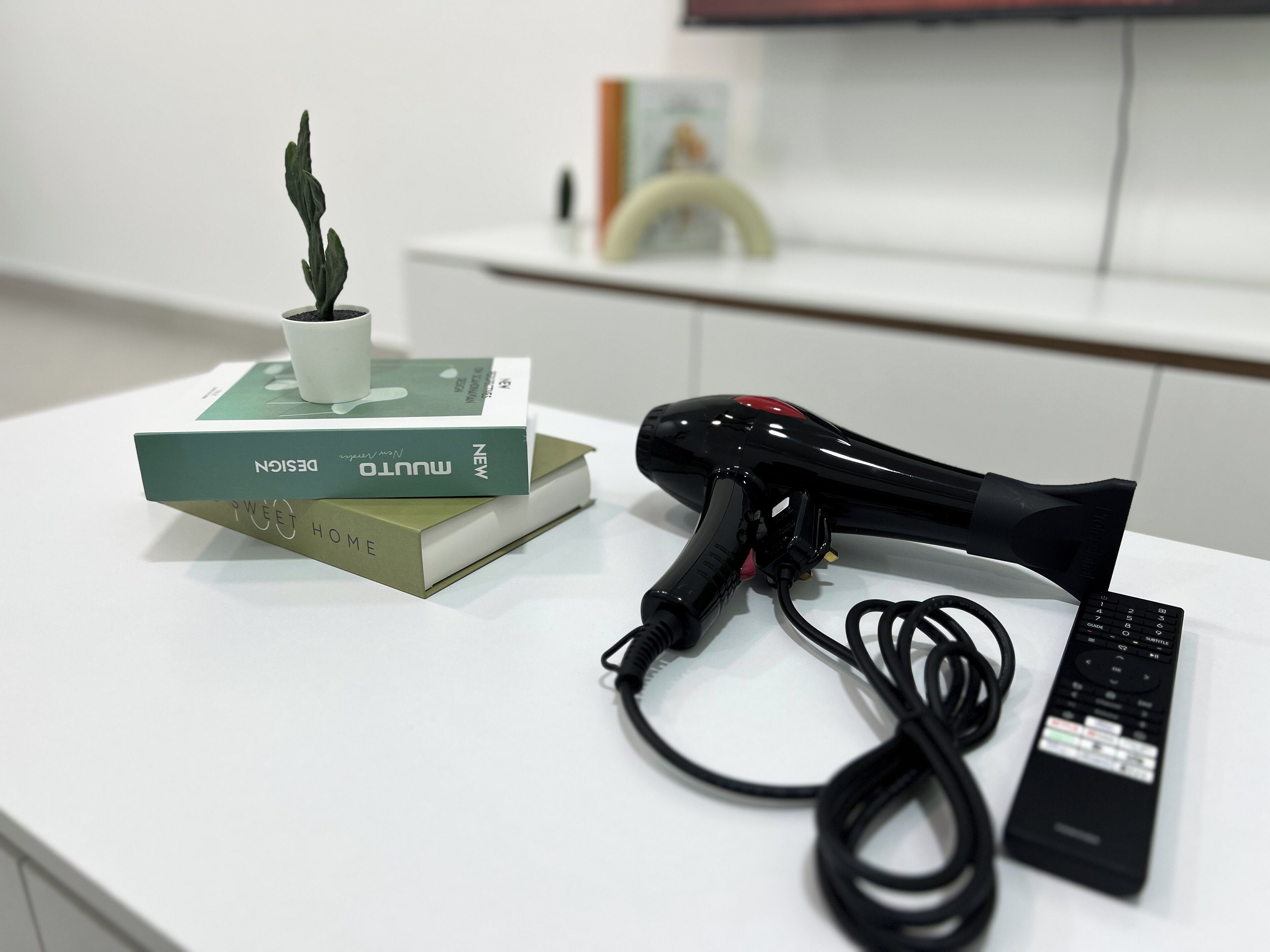 A white table with a black hairdryer, a remote control, and a stack of books.