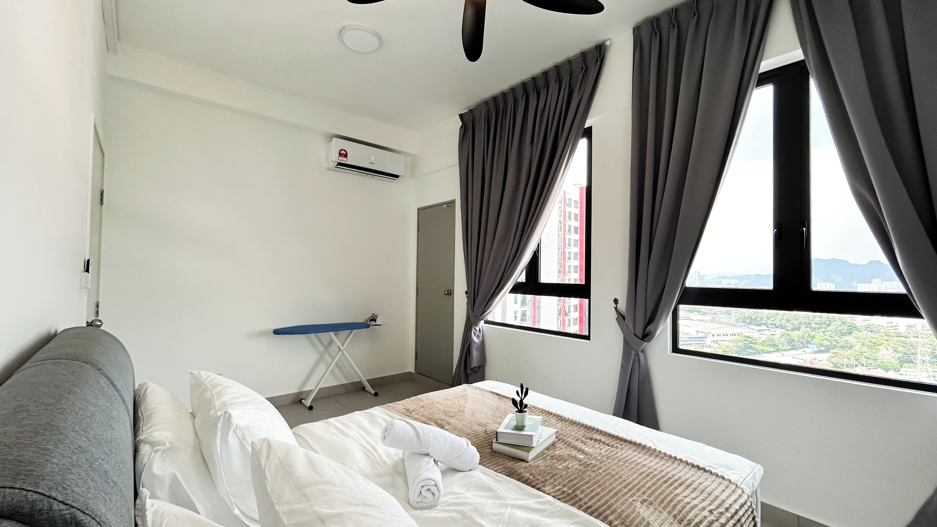 A hotel room with a bed, a window with curtains, an ironing board, and a wall-mounted air conditioner.