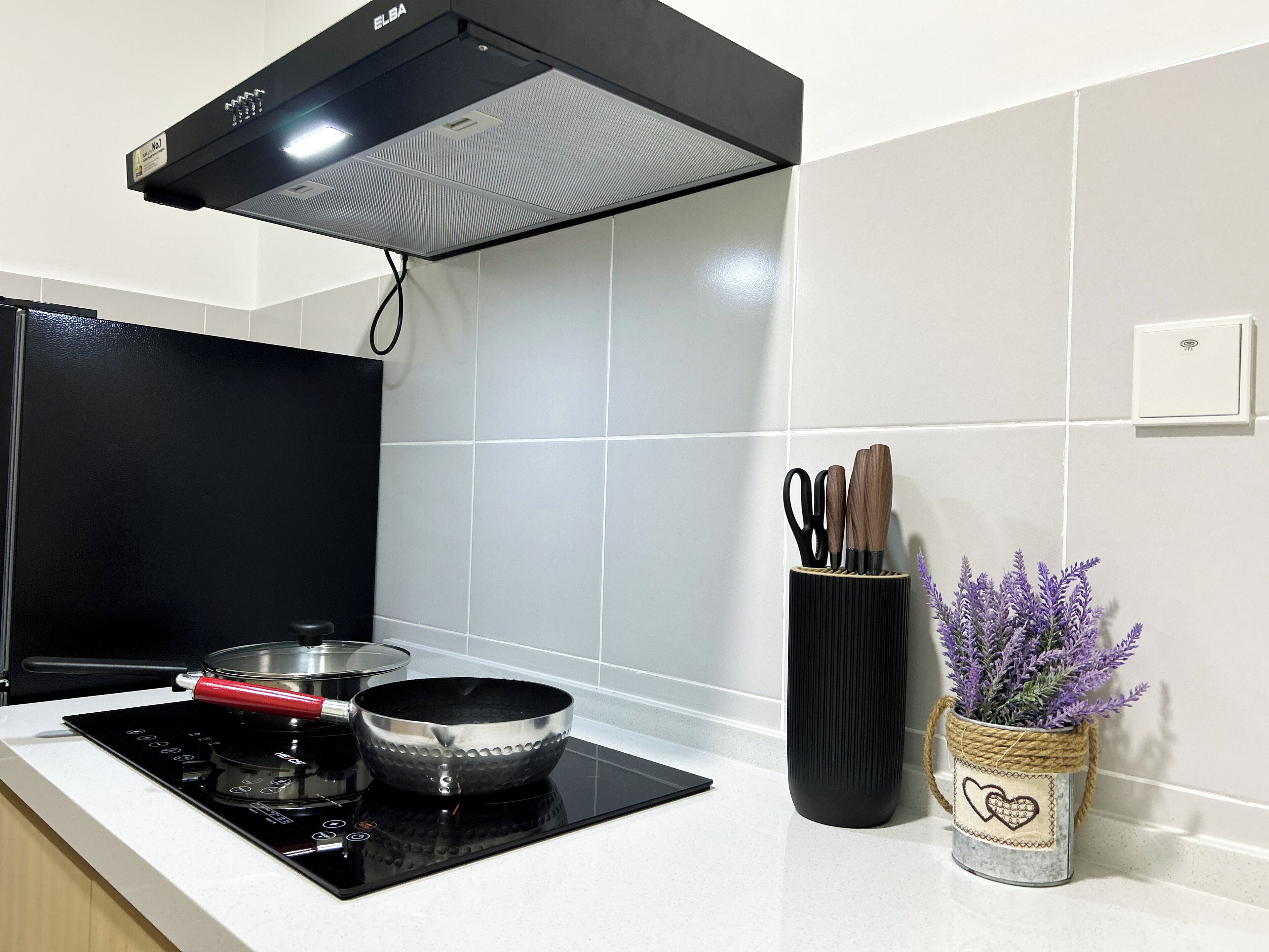 A modern kitchen with a black induction cooktop, a stainless steel pan, and a black pot on the cooktop. There is a black pot lid on the counter. A black pot is on the stove. A black pot is on the counter.