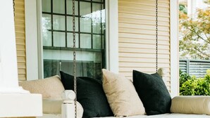 A porch with a white swing, a window with blinds, and a planter with flowers. - Luxe Farmhouse near Deep Ellum & Bishop Arts (Dallas)
