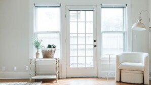 A room with a white door, two windows with blinds, a small table with a basket, a chair, and a lamp. - Luxe Farmhouse near Deep Ellum & Bishop Arts (Dallas)